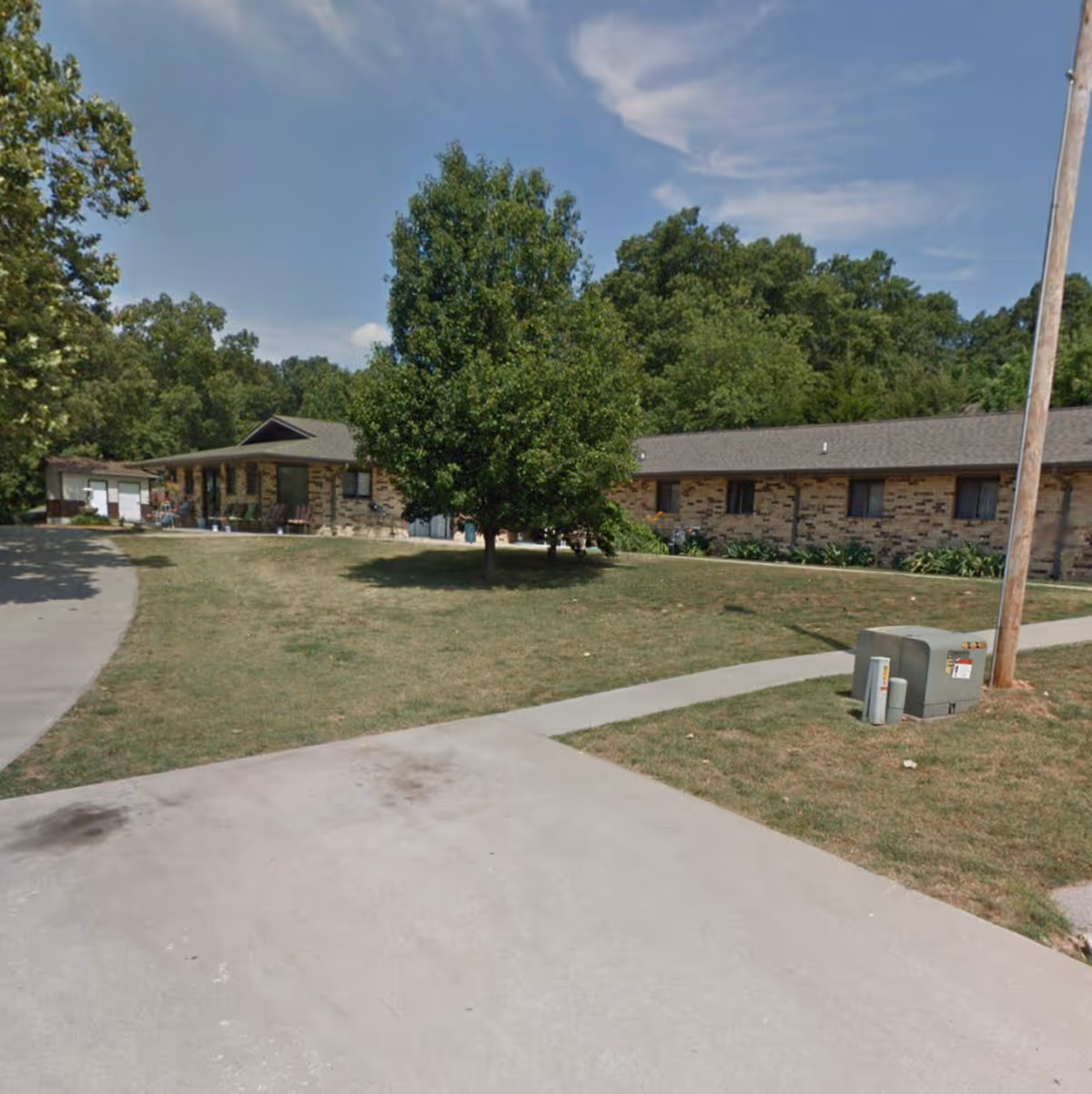 Exterior view of Cedar Ridge Care Center showing a single-story brick building with multiple windows, surrounded by green trees and a grassy lawn. A concrete driveway and sidewalk are visible in the foreground under a partly cloudy sky.