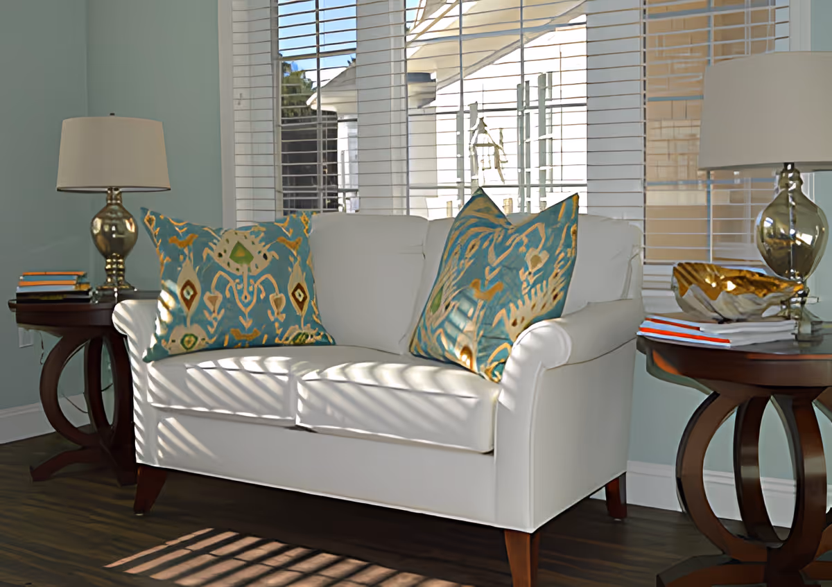 A bright living room area with a white loveseat adorned with two decorative blue and gold patterned pillows. On either side of the loveseat are dark wooden side tables, each holding a silver lamp with a white lampshade. One side table also has a stack of books, while the other has a decorative gold bowl and some papers. Behind the loveseat is a large window with white blinds partially open, allowing sunlight to cast shadows on the furniture and floor.