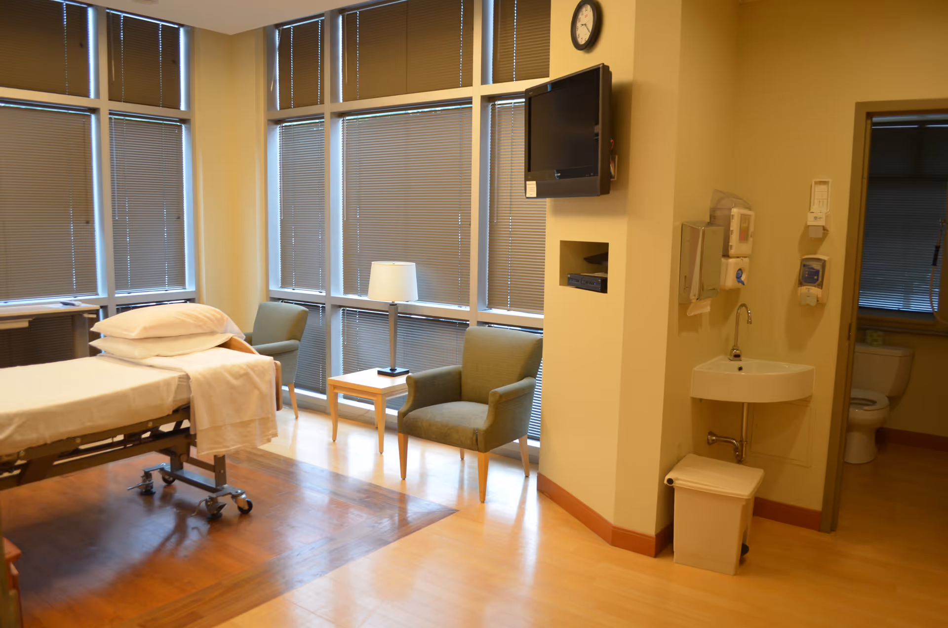 Hospital-style patient room with a bed, two chairs, a side table lamp, and wall TV.