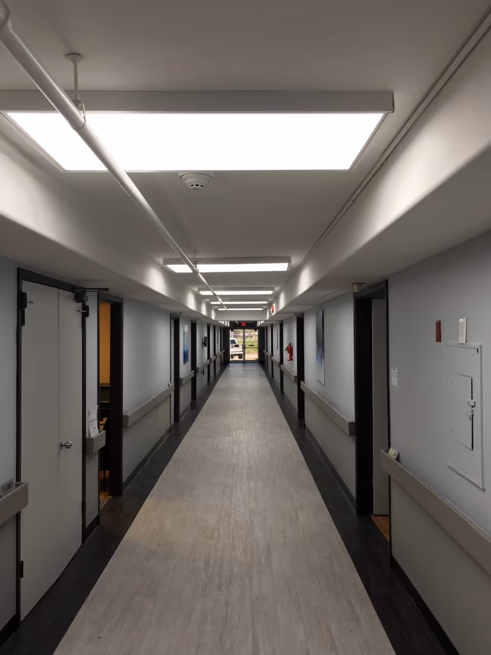A long, well-lit hallway in a senior living facility with light-colored flooring and gray walls. Doors line both sides of the corridor, and handrails run along the walls. Bright ceiling lights illuminate the space, and a window at the far end shows an outdoor view.