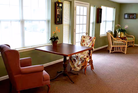 A cozy interior room with a wooden table and two chairs, one upholstered in a floral fabric and the other in a solid color. The room has green walls, multiple windows with white blinds, and a glass door. In the background, there are two wicker chairs with cushions and a small table with a plant on it.