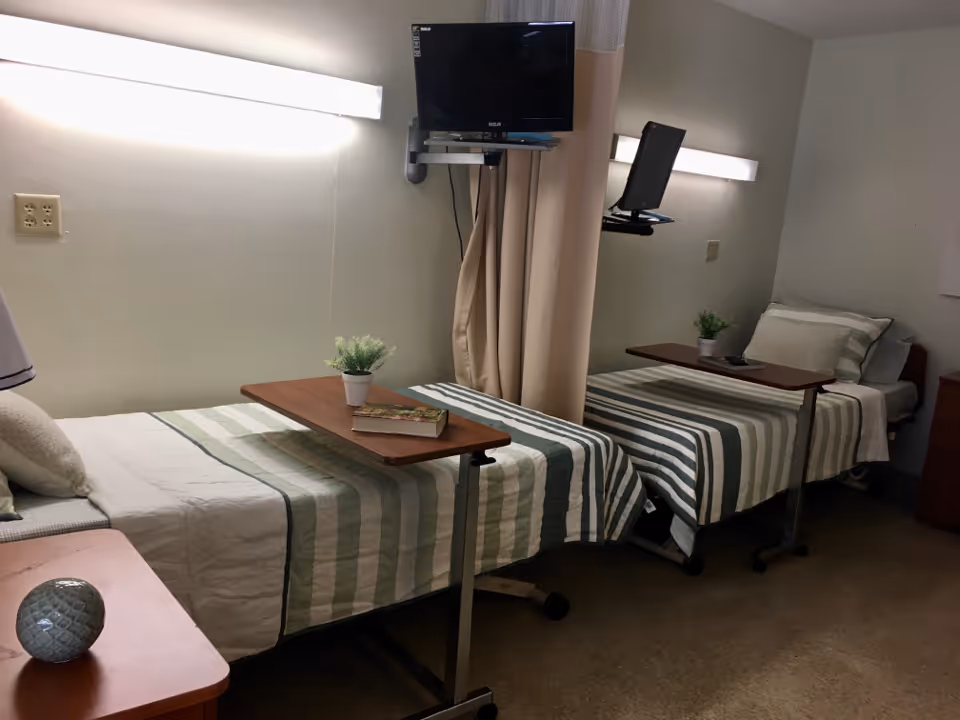 Two neatly made twin beds with green-and-white striped bedding, overbed tables, a wall-mounted TV, and a privacy curtain in a shared senior facility room.