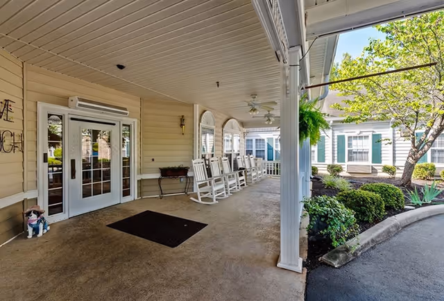 Covered front entrance/porch with a row of white rocking chairs, double glass doors, and landscaped beds beside the driveway.