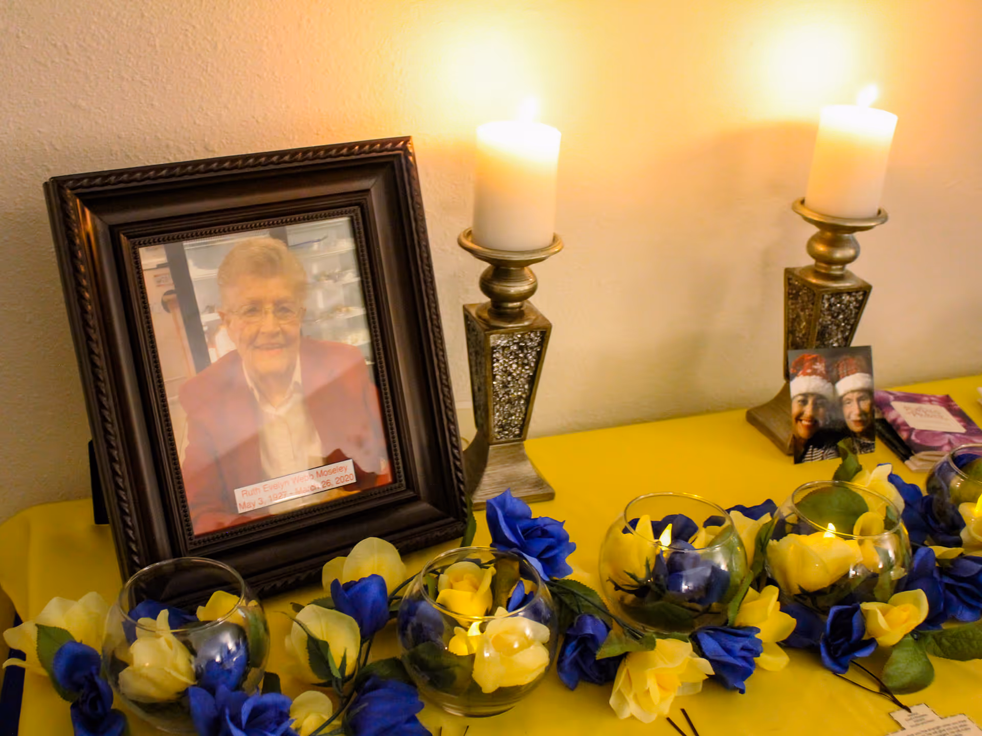 Memorial table with framed elderly woman's photo, lit candles, and blue and yellow flowers.
