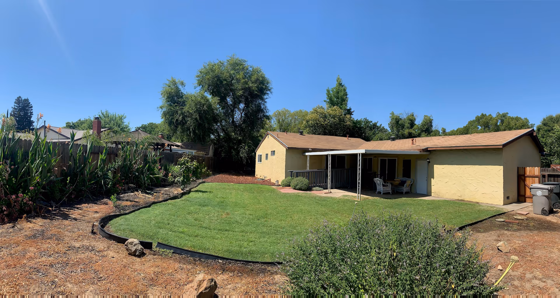Backyard of a single-story yellow house with a covered patio, a green lawn, planted borders and a wooden fence under a clear blue sky.