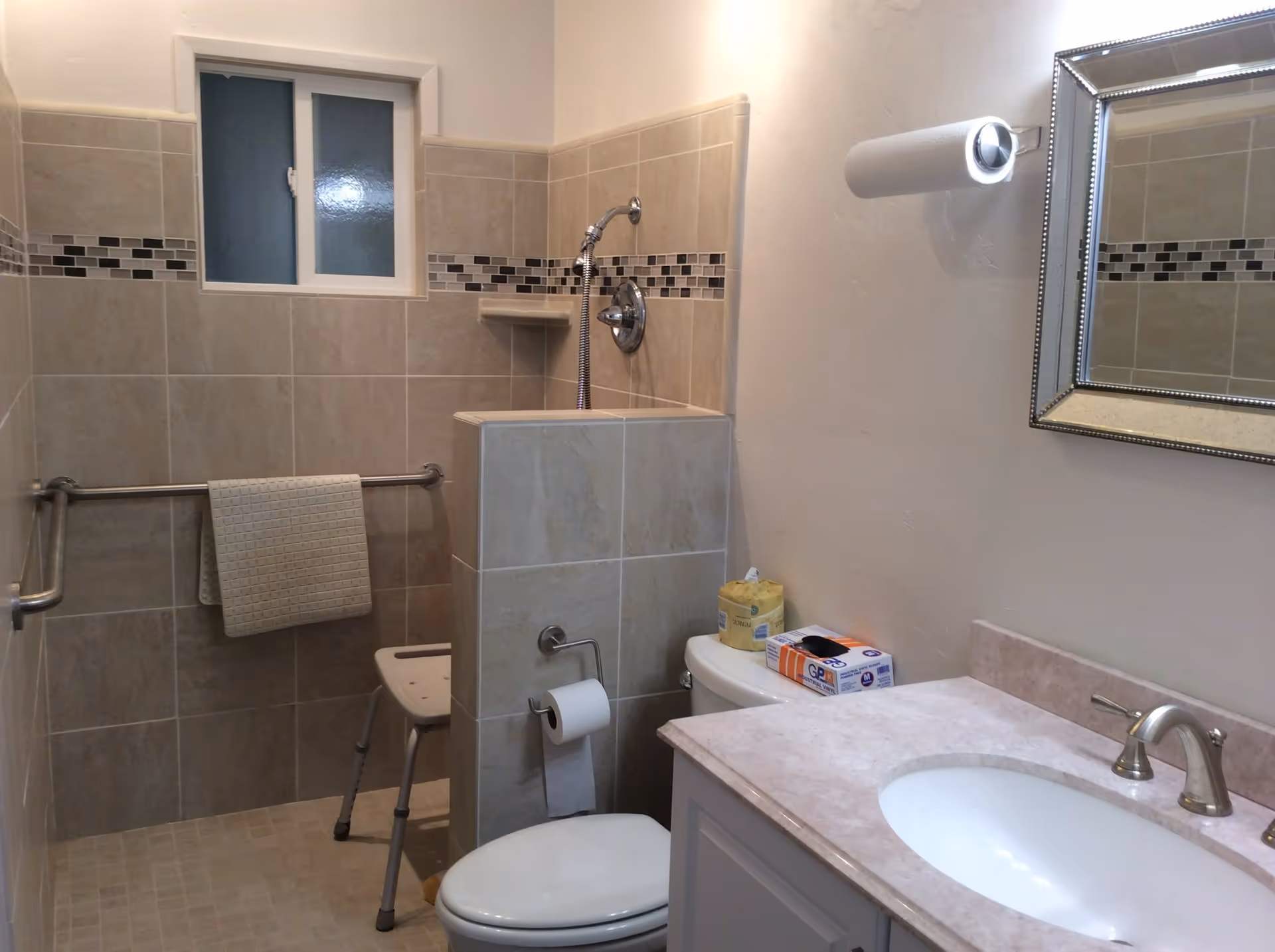 A bathroom with beige tiled walls and floor, featuring a walk-in shower with a handheld showerhead and a shower chair. There is a grab bar with a towel hanging on it, a toilet with a roll of toilet paper on the holder, and a countertop with a sink and faucet. A mirror is mounted on the wall above the sink, and a paper towel holder is attached to the wall.