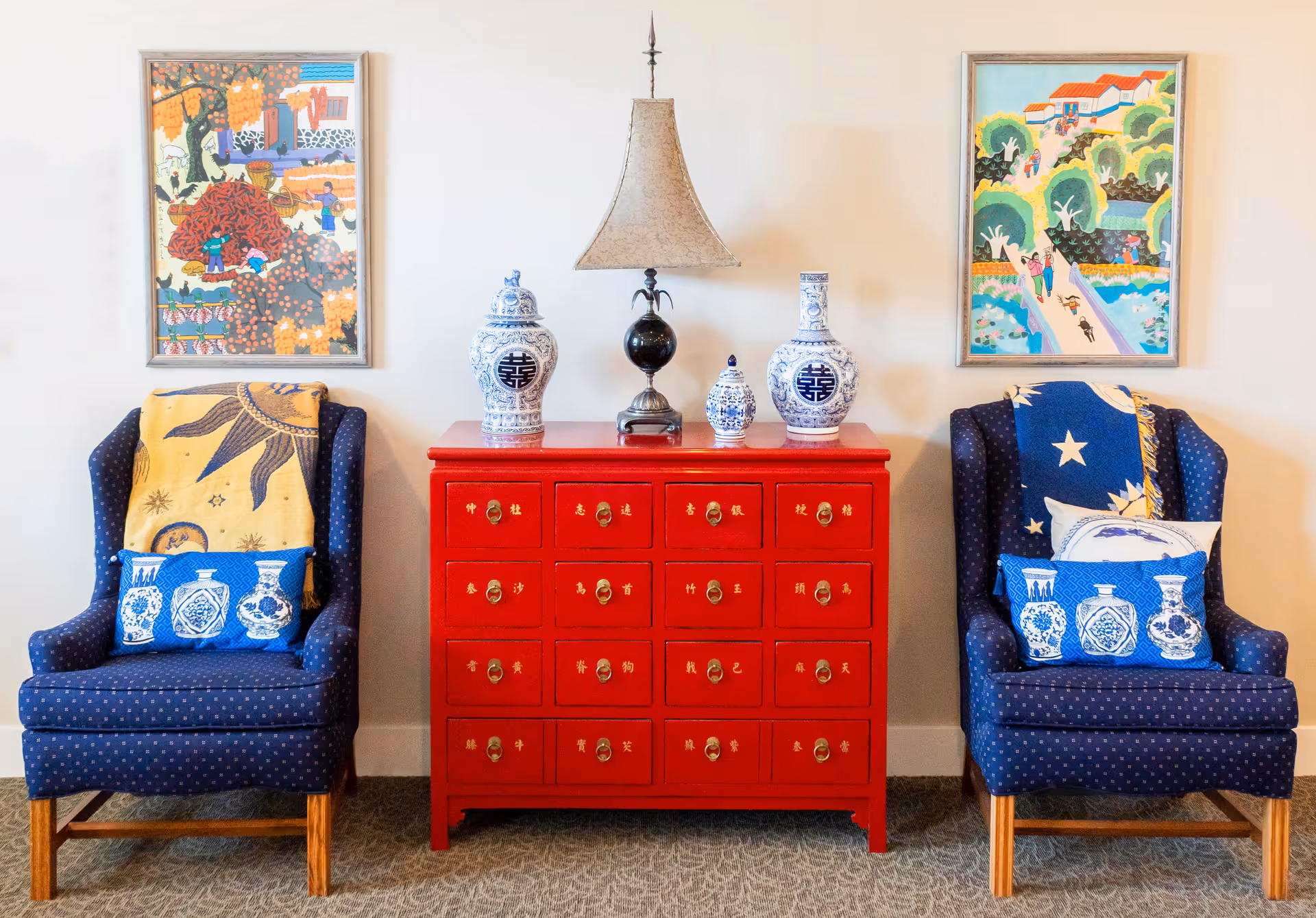 Two blue upholstered armchairs flank a red chest topped with blue-and-white vases and a lamp, with framed colorful paintings on the wall behind.