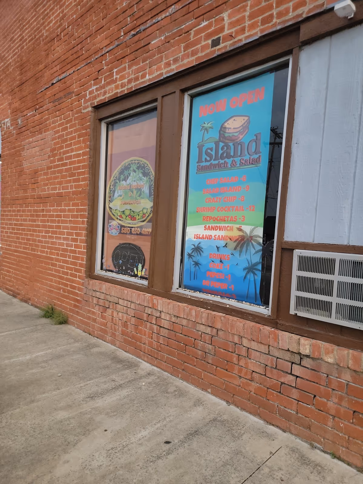 Brick storefront exterior with two windows displaying colorful posters for Island Sandwich & Salad.