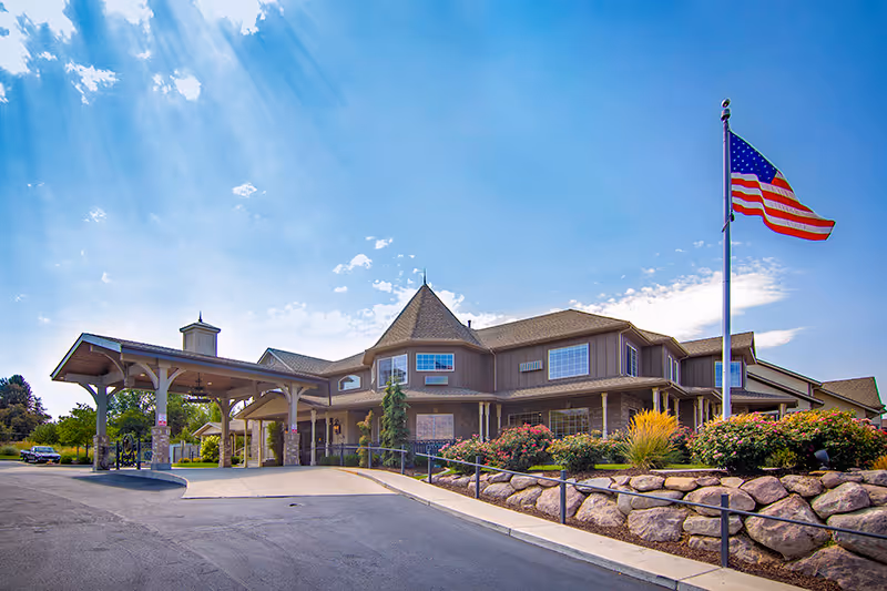 Front exterior of a two-story assisted living building with a covered entrance, landscaped rock beds, and an American flag flying on a flagpole.