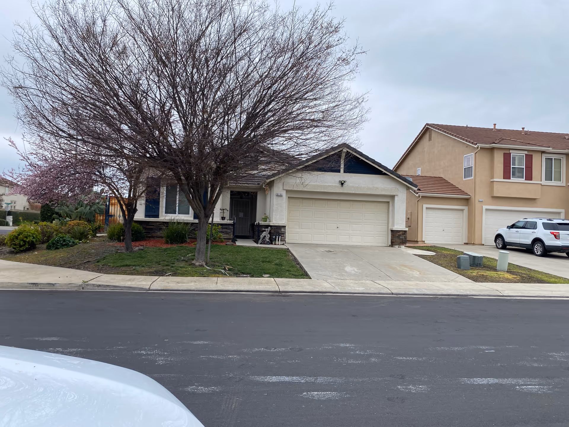 Front exterior view of a single-story residential house with a two-car garage, a leafless tree in the front yard, and a concrete driveway. The house has light-colored walls with dark trim and a small porch area. Adjacent to the house is a two-story home with a beige exterior and a white SUV parked in the driveway. The sky is overcast.
