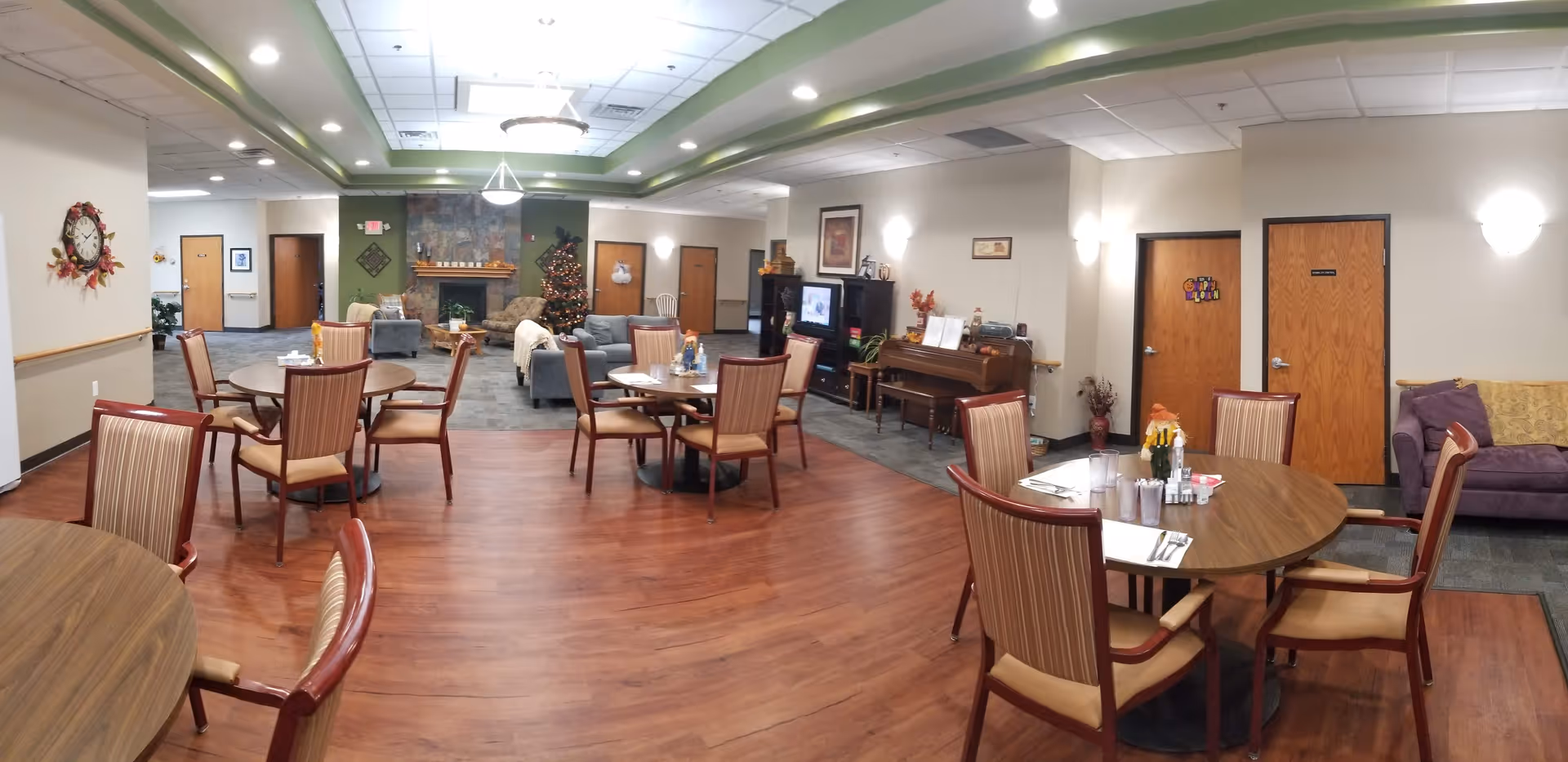 A spacious senior living common area with several round wooden tables and chairs arranged on a wood floor. In the background, there is a cozy sitting area with sofas, a decorated Christmas tree, and a stone fireplace. The walls are light-colored with some framed artwork and a piano along one side. The ceiling has recessed lighting and a green accent border.
