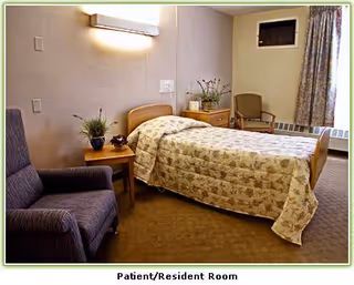 Patient room with single bed, quilted bedspread, chairs, and bedside table.
