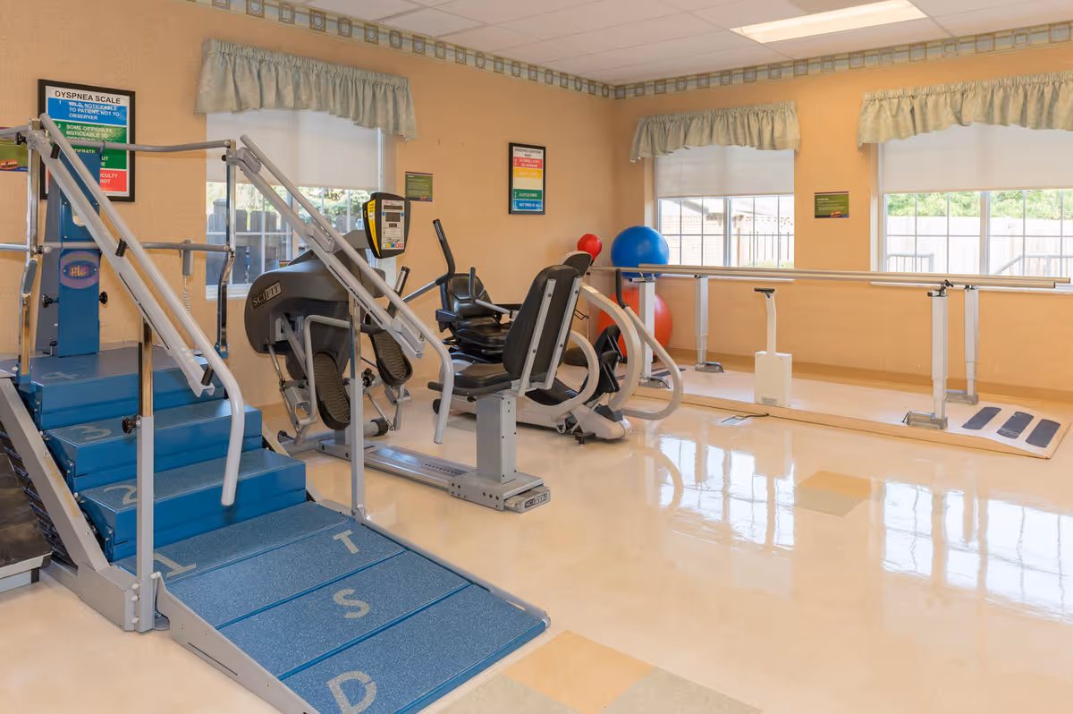 Indoor fitness room with exercise machines, parallel bars, and gym equipment in a senior living facility.