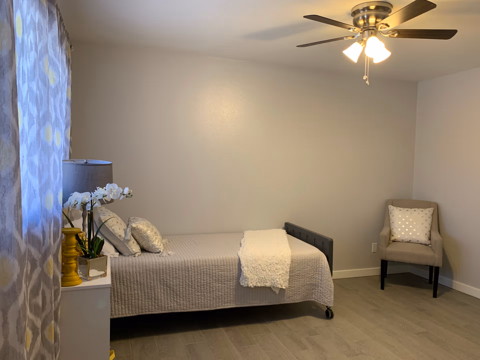 A simple bedroom with a single bed covered in a beige quilt and white throw blanket. There are decorative pillows on the bed. Next to the bed is a white nightstand with a yellow lamp and a white orchid plant. A beige armchair with a polka dot pillow is placed in the corner. The room has light-colored walls, wood flooring, a ceiling fan with lights, and patterned curtains on the window.