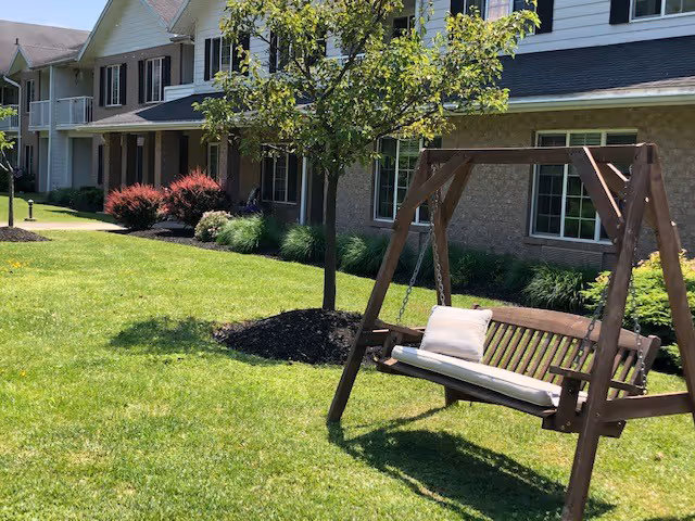 A grassy front lawn with a wooden porch swing and small tree in front of a senior living building's facade.