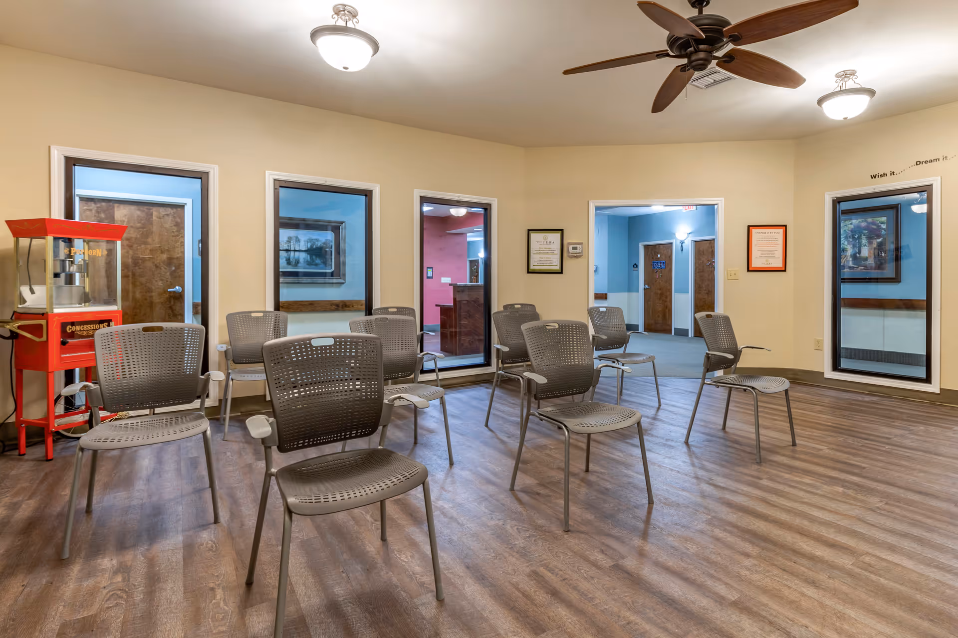 A room with several gray plastic chairs arranged in rows on a wood floor. The walls are beige with three large windows and a doorway leading to other rooms. There is a red popcorn machine in the corner and ceiling fans with lights above. Various framed pictures and certificates hang on the walls.