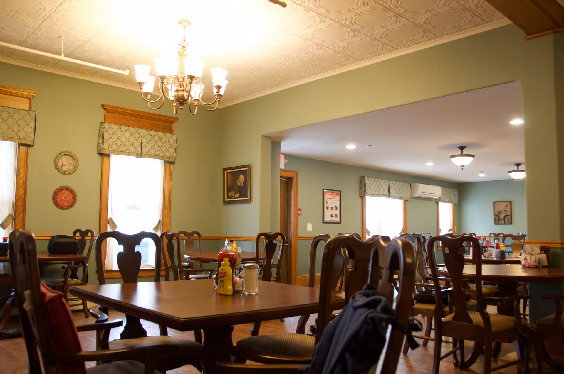 A dining room with multiple wooden tables and chairs arranged neatly. The walls are painted green with wooden trim around the windows and doorways. There are decorative plates and framed pictures on the walls. A chandelier with multiple lights hangs from the ceiling, and windows have patterned valances. Condiments such as ketchup and mustard are placed on the tables.