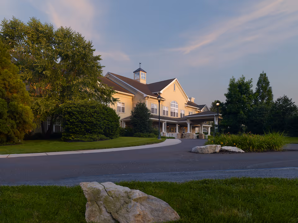 Front exterior of a two-story senior living facility with a portico entrance, driveway and landscaped lawns and trees at dusk.