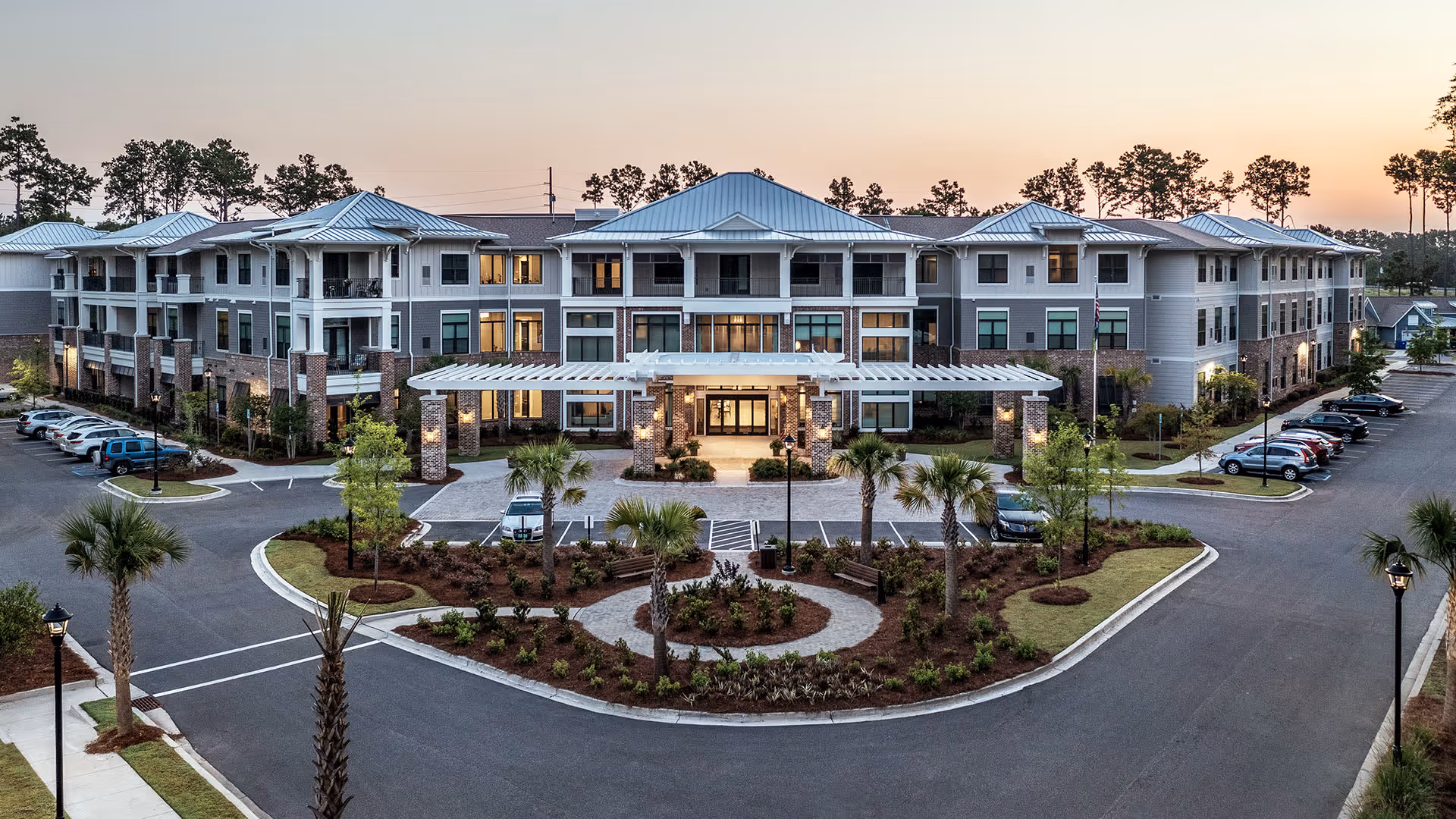 Exterior view of The Artisan Mount Pleasant senior living facility at dusk, showing a large three-story building with a covered entrance, landscaped roundabout with palm trees and benches, and parked cars on either side.