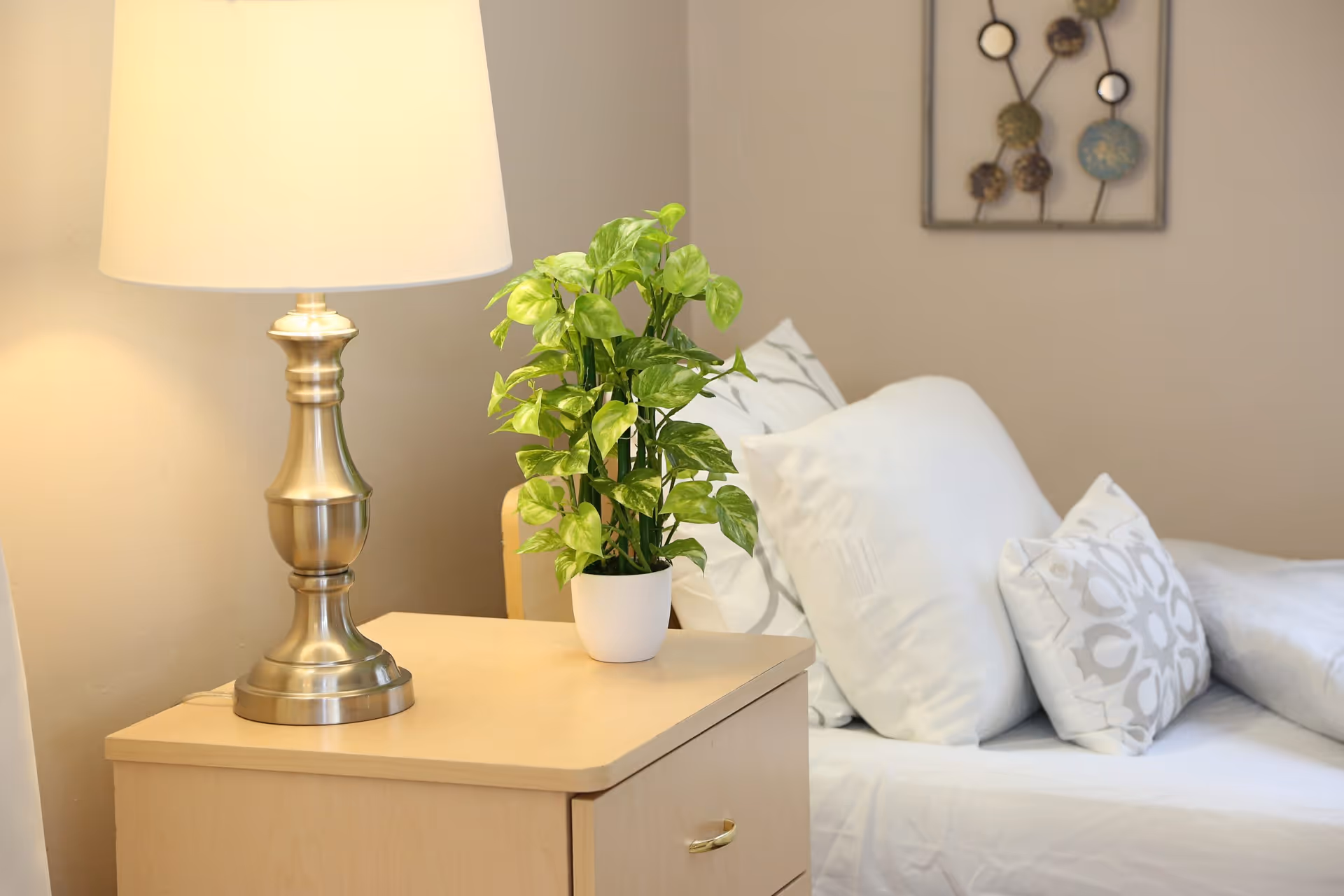 A bedside table with a lamp and potted plant next to a bed with white pillows and decorative wall art.