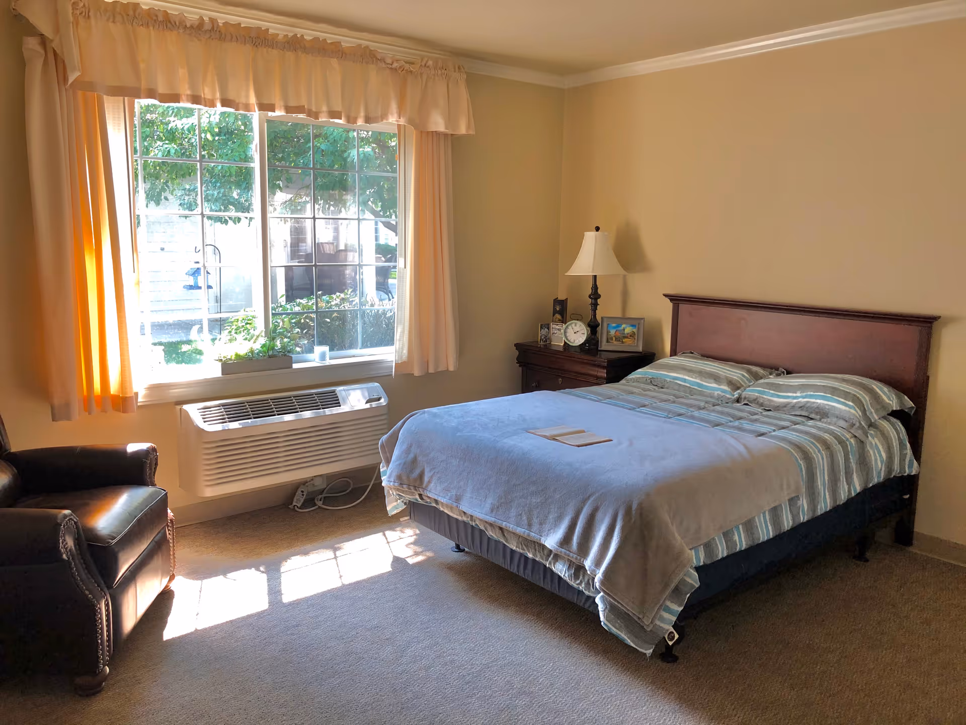 Sunlit bedroom with a large bed, nightstand, lamp, window, and leather chair.