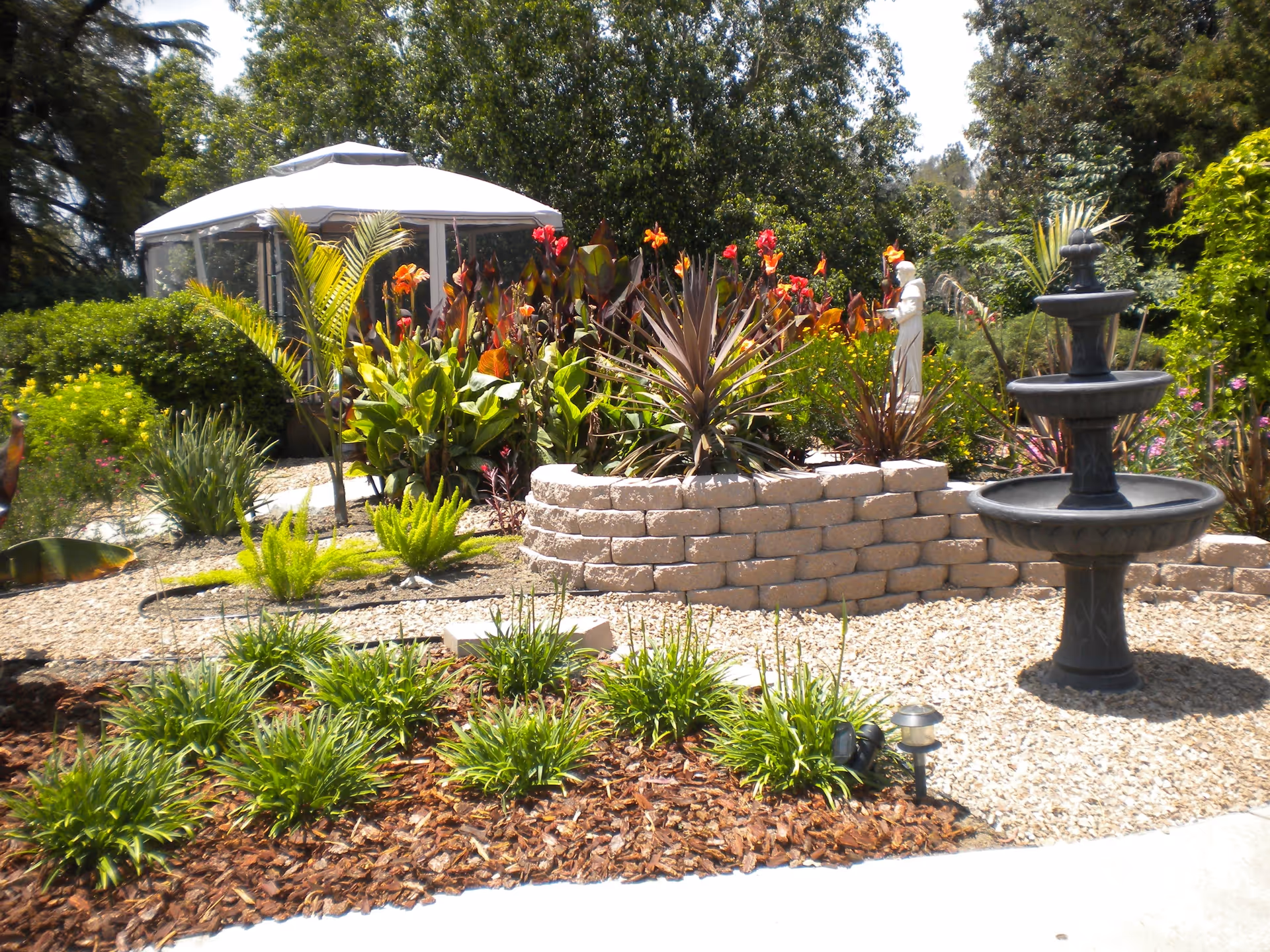 A sunny outdoor garden with a tiered fountain, stone planter, and gazebo.