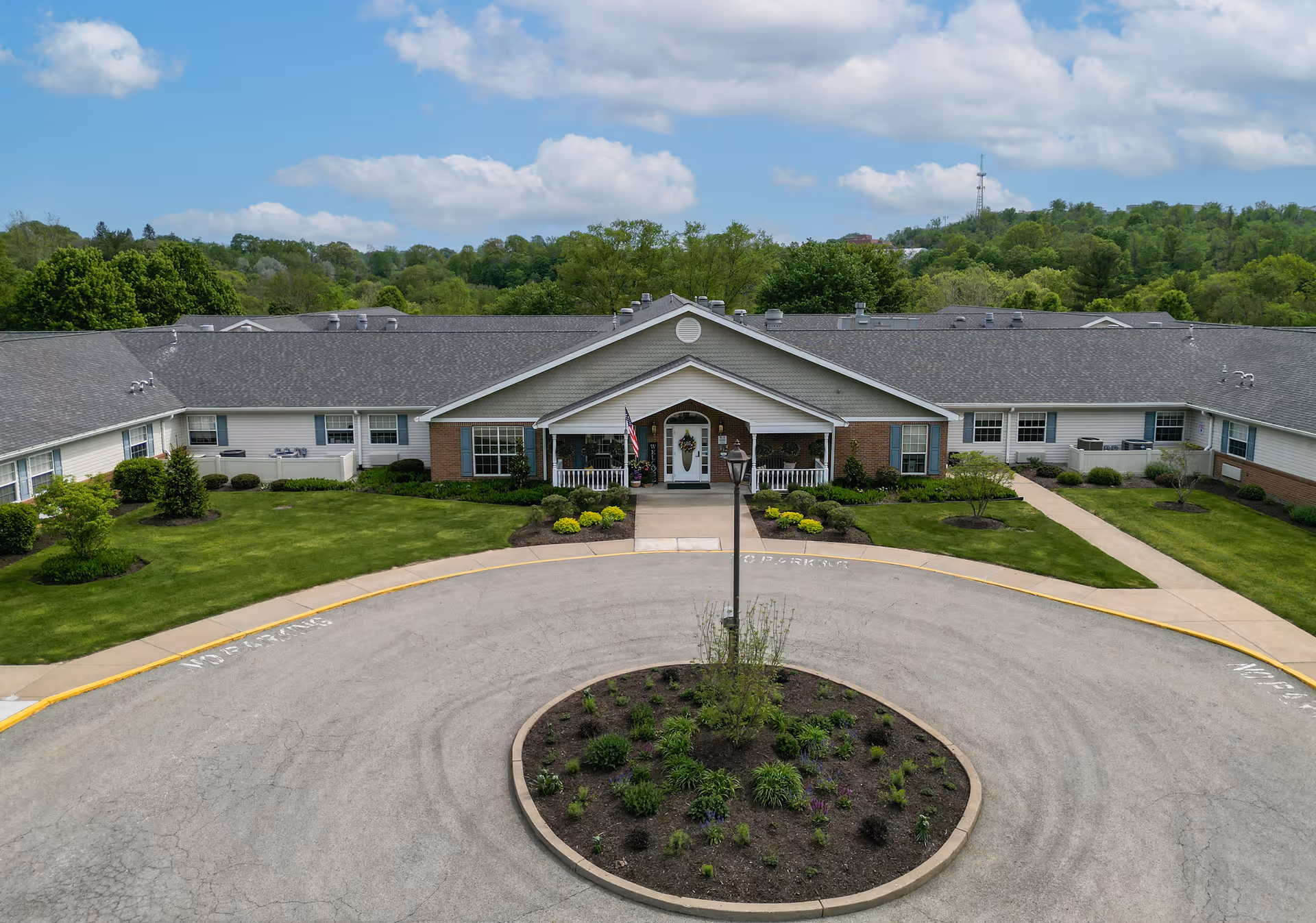 Front exterior view of Arden Courts - ProMedica Memory Care Community in Jefferson Hills, showing a single-story building with a covered entrance, a circular driveway with a landscaped center island, green lawns, and trees in the background under a partly cloudy sky.