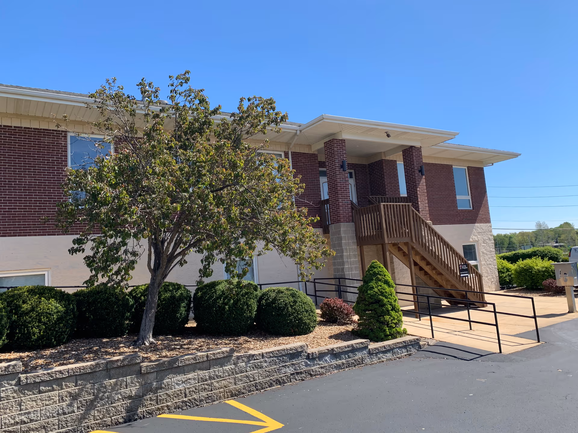 Exterior view of a brick senior living facility with stairs, landscaping, and a clear blue sky.