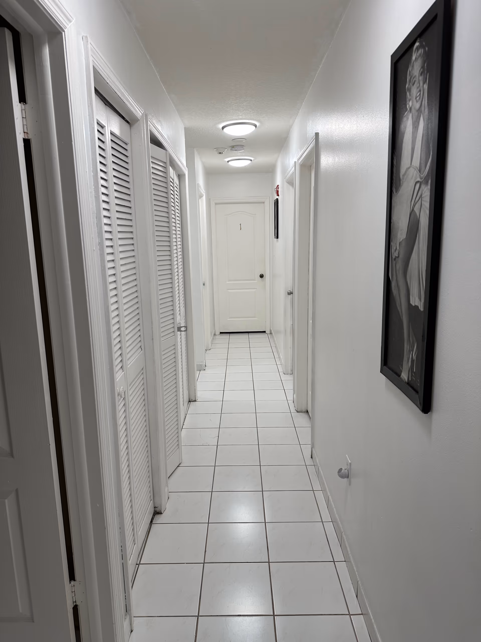 Long white hallway with tiled floor, doors, and framed art along the walls.