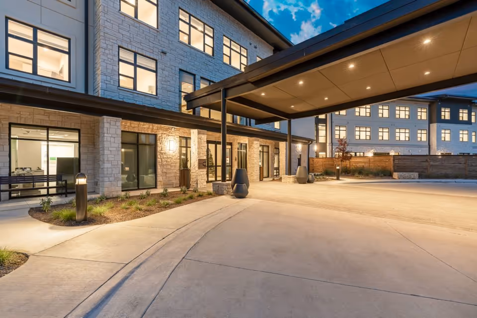 Evening view of the exterior entrance of a senior living facility named The Reserve at Lake Austin, featuring stone and stucco walls, large windows, a covered drop-off area, and well-lit pathways with landscaping.