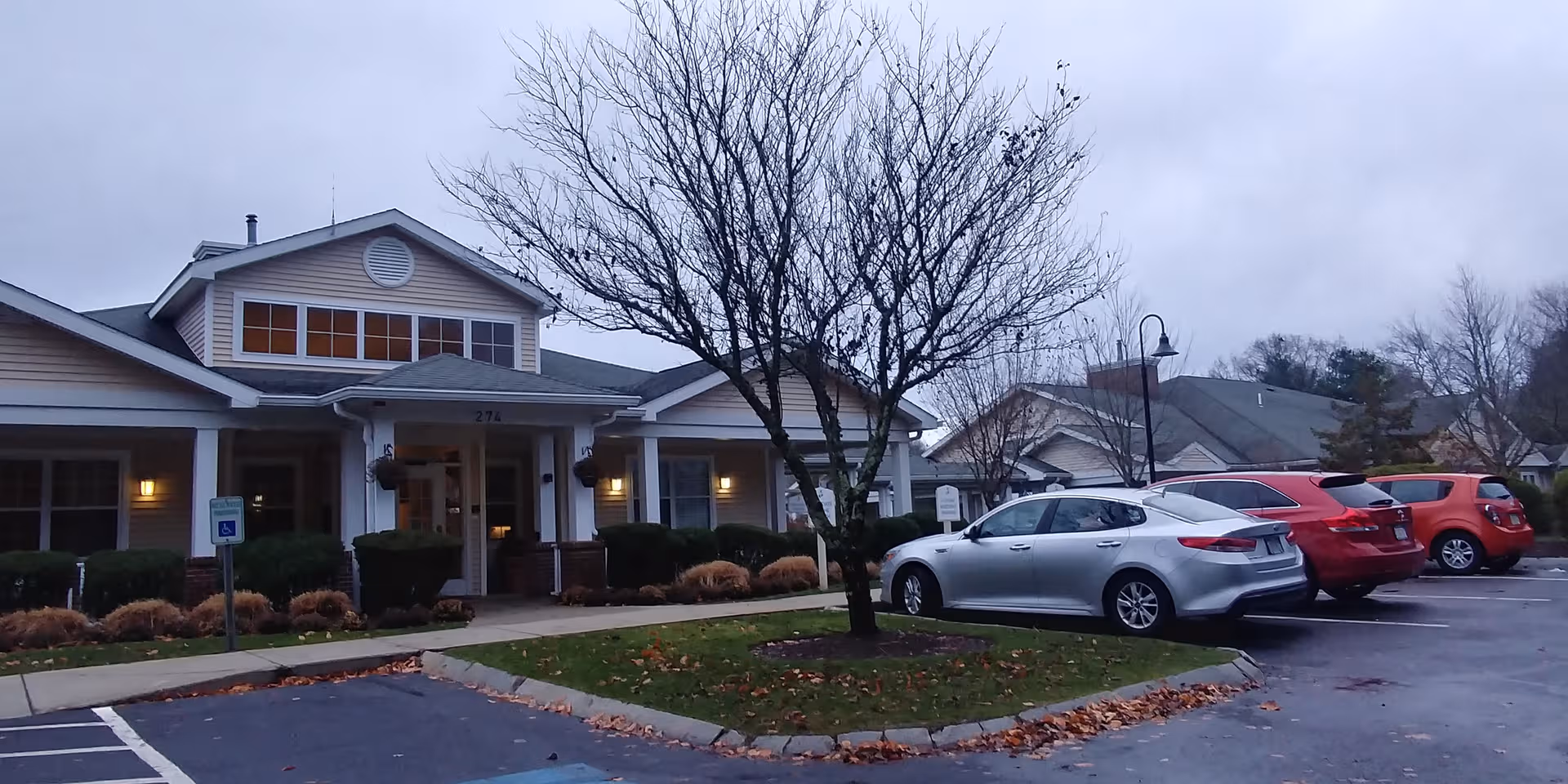 Senior living facility with front entrance, parking lot, and leafless tree.
