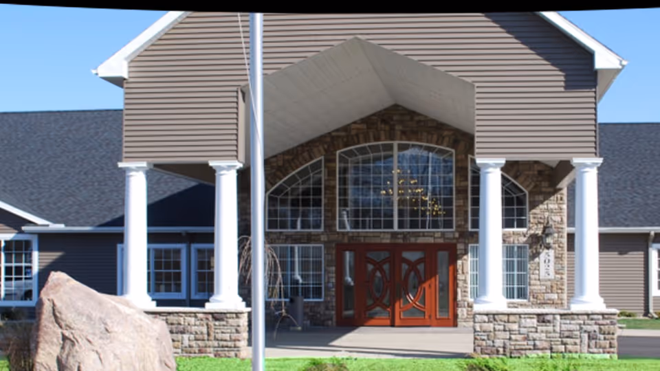 Front exterior view of a building with stone and siding facade, featuring large windows and double wooden doors under a covered entrance supported by white columns.