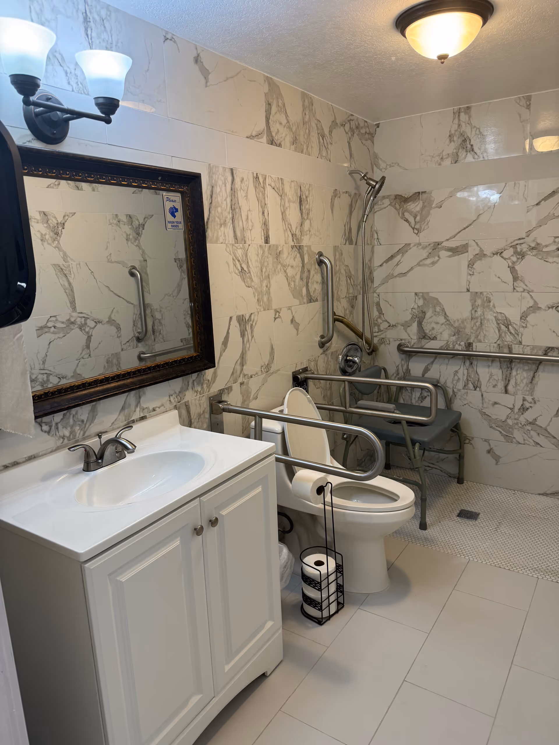 Accessible bathroom with marble walls, sink vanity, mirror, toilet, shower, and grab bars.