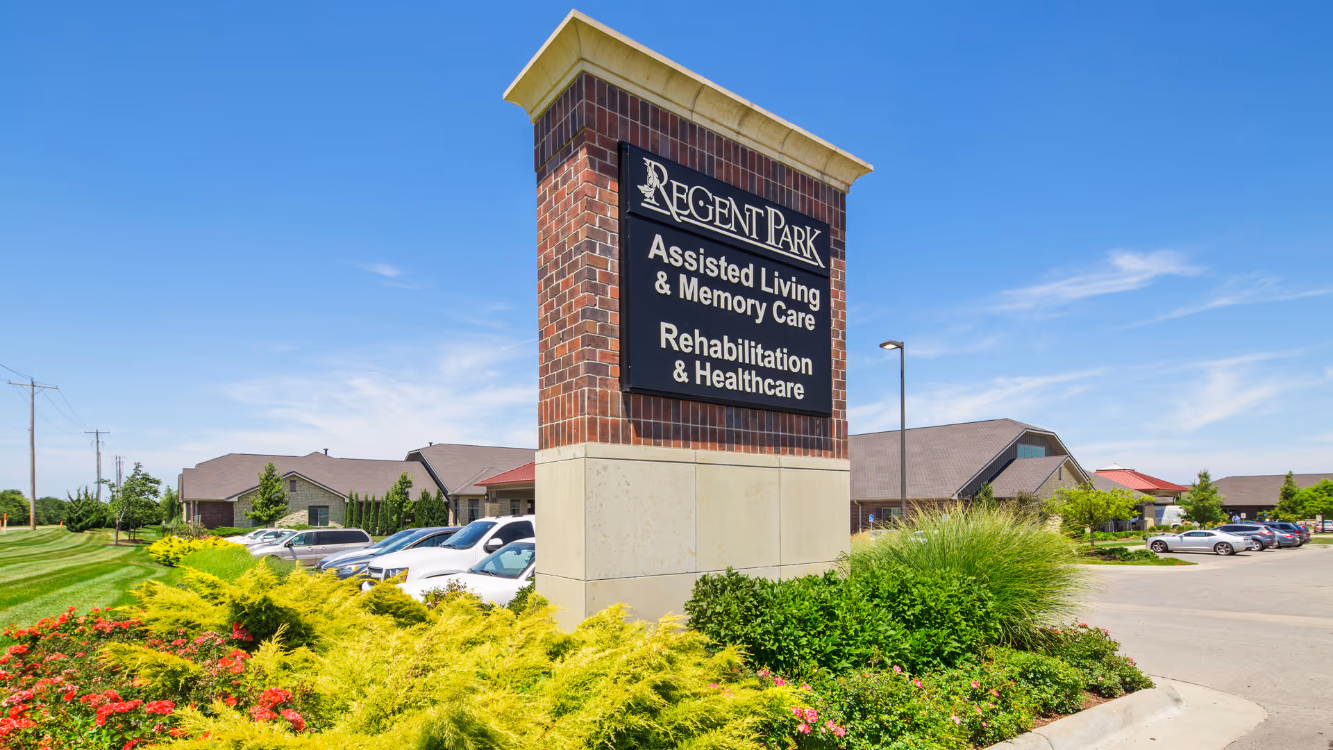 Exterior view of a brick entrance sign reading "Regent Park Assisted Living & Memory Care" with landscaped grounds, parked cars, and the facility building behind.