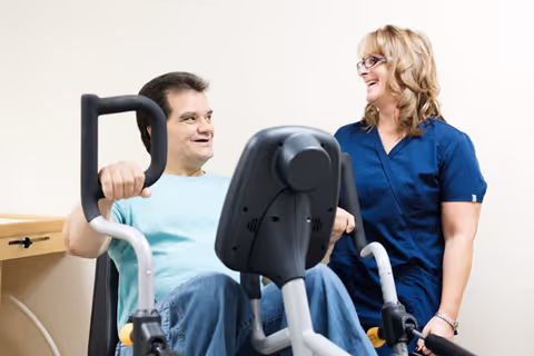 A man using a seated exercise machine while a woman in blue scrubs stands beside him, smiling and engaging with him in a light-colored room.