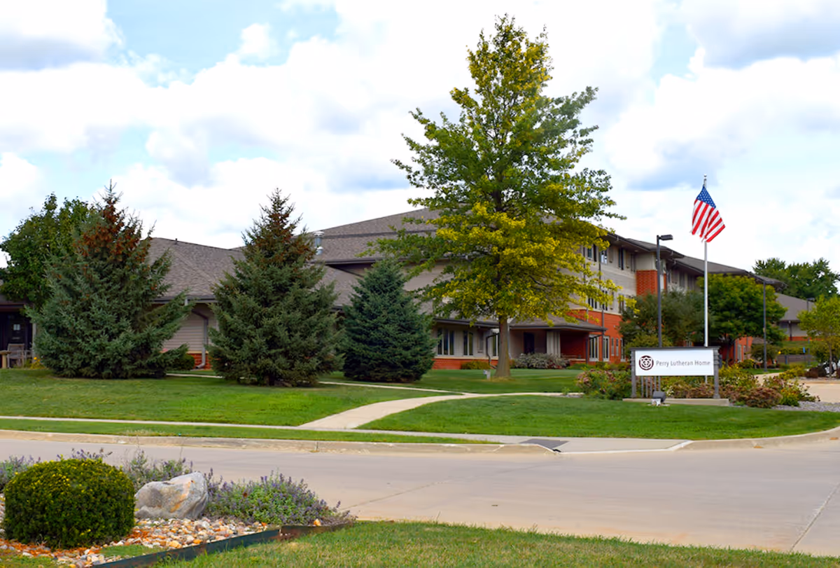 Exterior view of the Spring Valley Campus of Perry Lutheran Homes building with a well-maintained lawn, several trees, a sidewalk, an American flag on a flagpole, and a sign displaying the facility's name.
