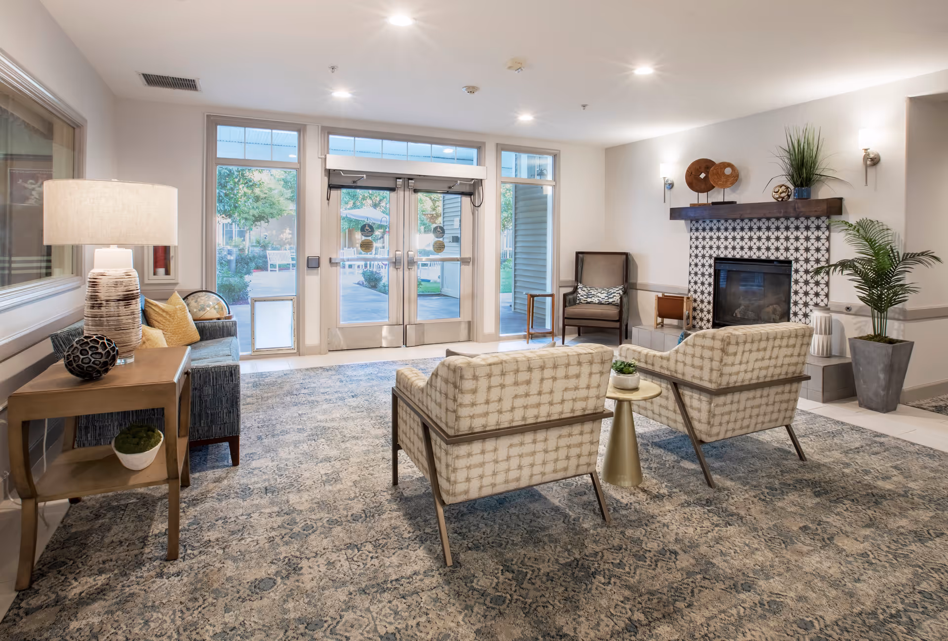 A bright and inviting common area with two patterned armchairs facing a decorative fireplace with a tiled surround. There is a small round table between the chairs with a small plant on it. To the left, there is a wooden side table with a lamp and decorative items, next to a cushioned bench with pillows. Large glass double doors and windows let in natural light and provide a view of an outdoor garden area. The room is decorated with plants and wall-mounted lights.