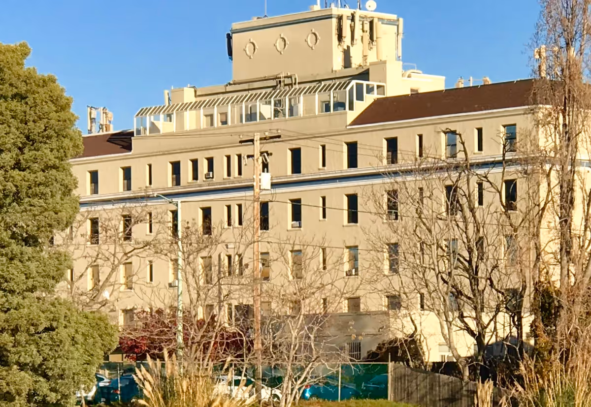 Exterior view of a multi-story beige hospital building with numerous windows, surrounded by trees and some parked cars, under a clear blue sky.