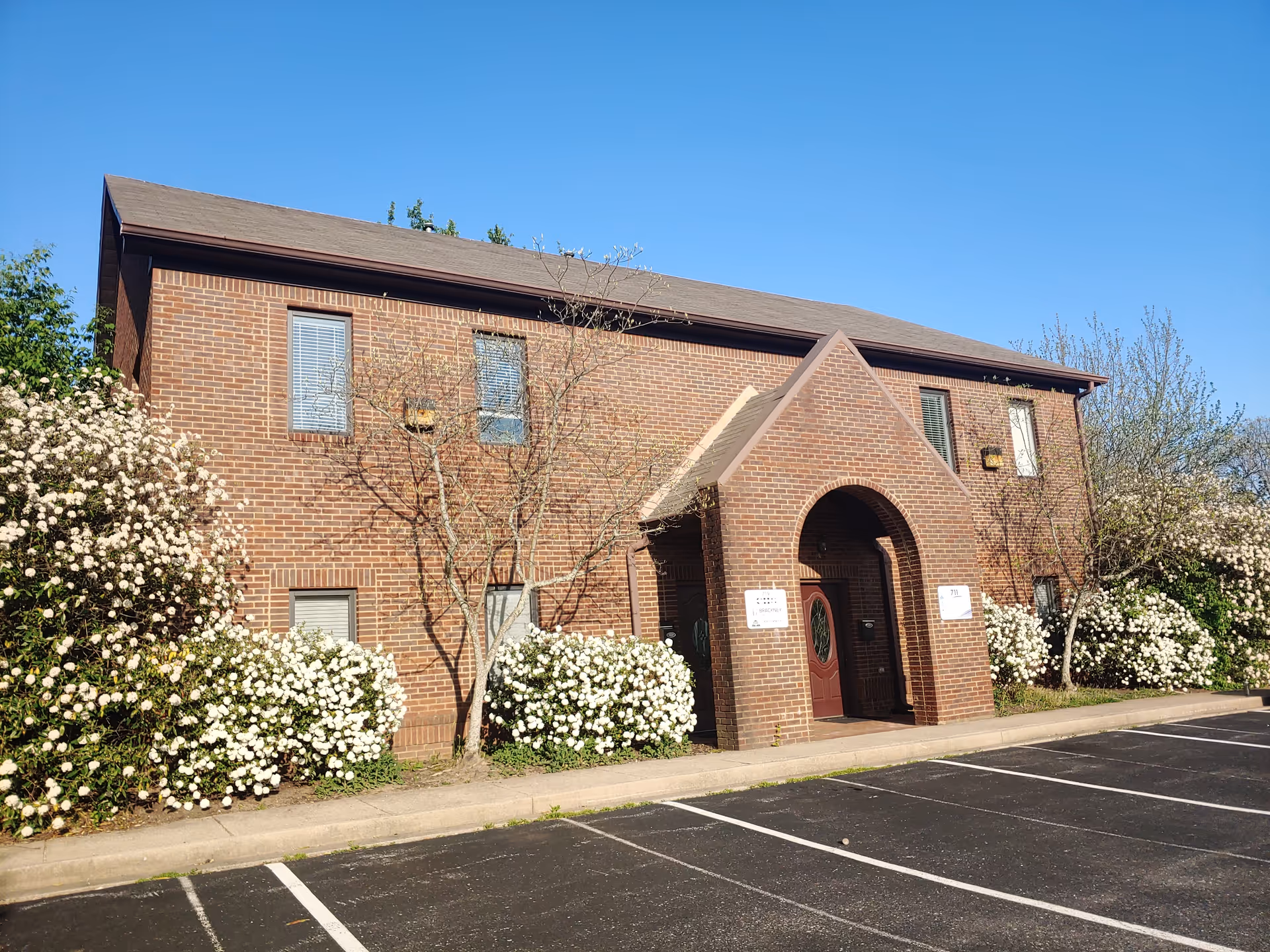 Brick two-story building with arched entry, blooming shrubs, and a paved parking lot under blue sky.