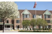 Brick senior living facility exterior with green peaked dormers, trees, and an American flag.