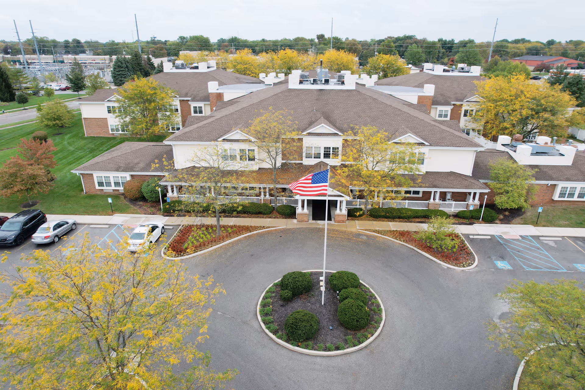 Aerial view of Carriage Court Senior Living building with a circular driveway featuring a flagpole with an American flag in the center. The building has a brown roof, white walls, and multiple windows, surrounded by trees with autumn foliage and parked cars.