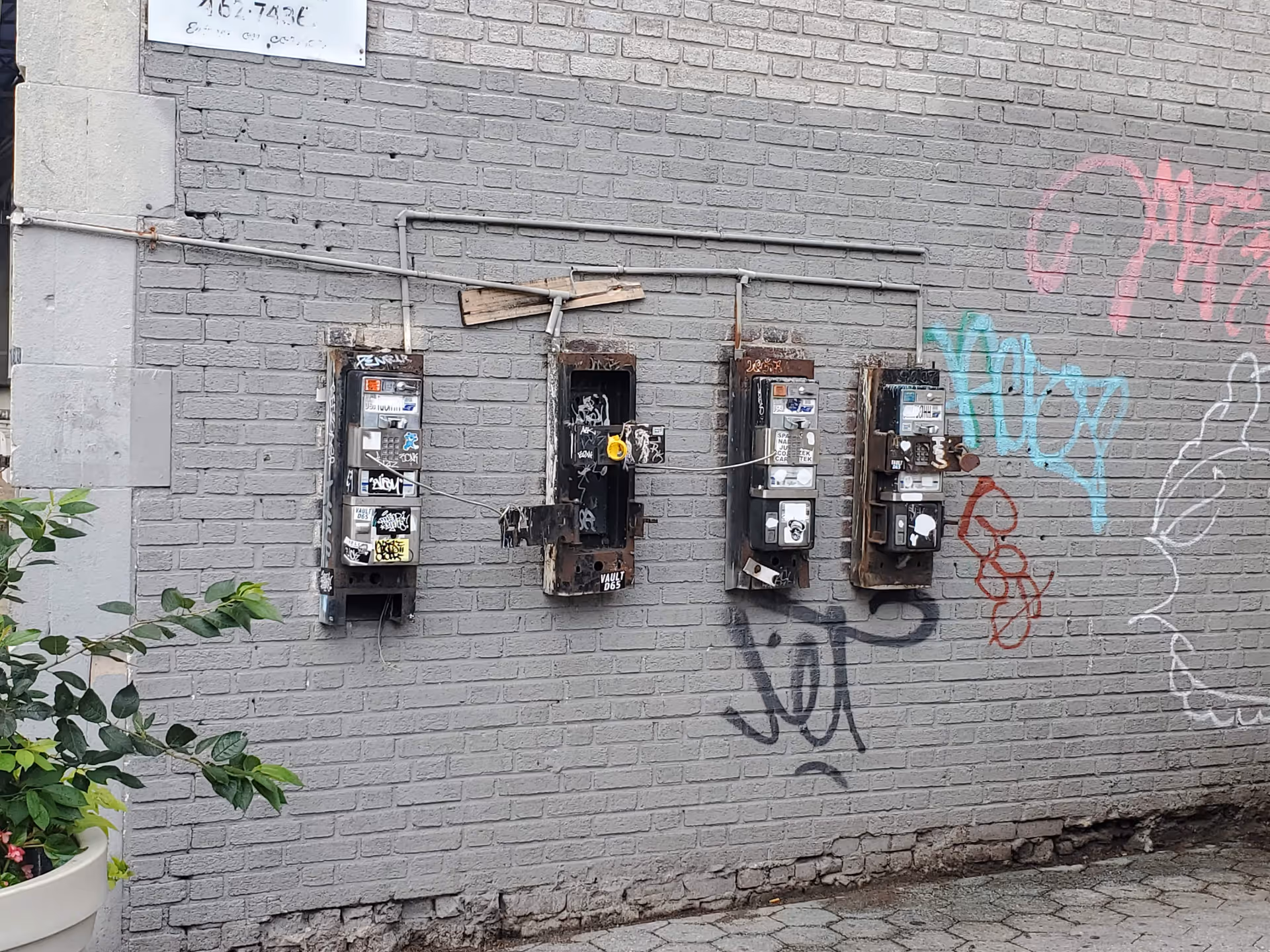 Weathered brick exterior wall featuring three old payphones, graffiti, and a potted plant.