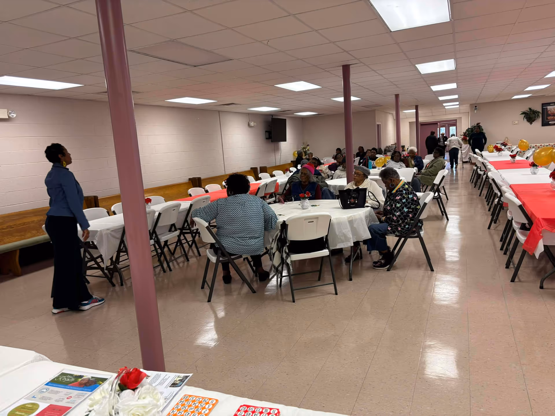Senior living facility dining hall with residents seated at round tables for a gathering.