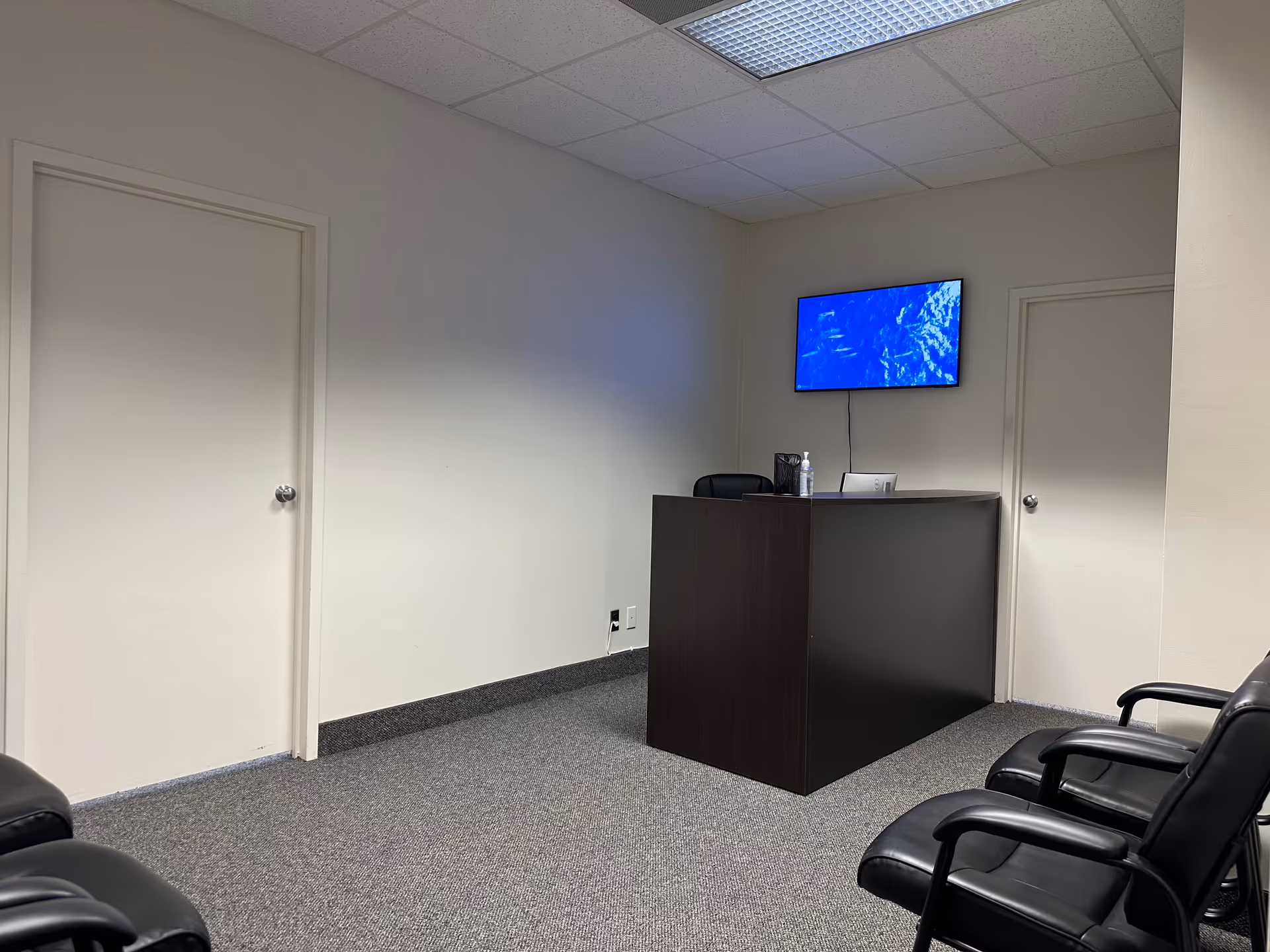 Small reception area featuring a dark desk, wall-mounted monitor, and row of black chairs.