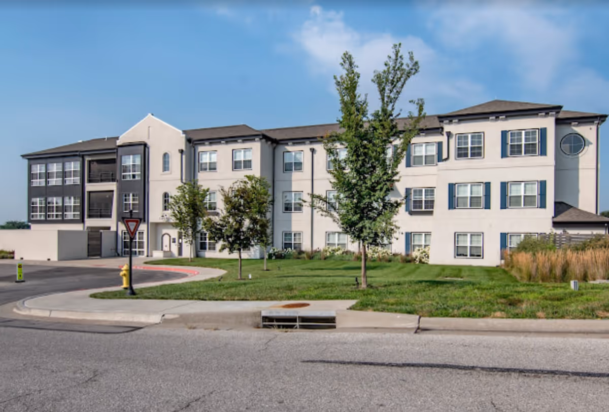 Three-story light-colored senior living building with multiple windows and balconies, a grassy lawn and trees in front and a circular driveway.