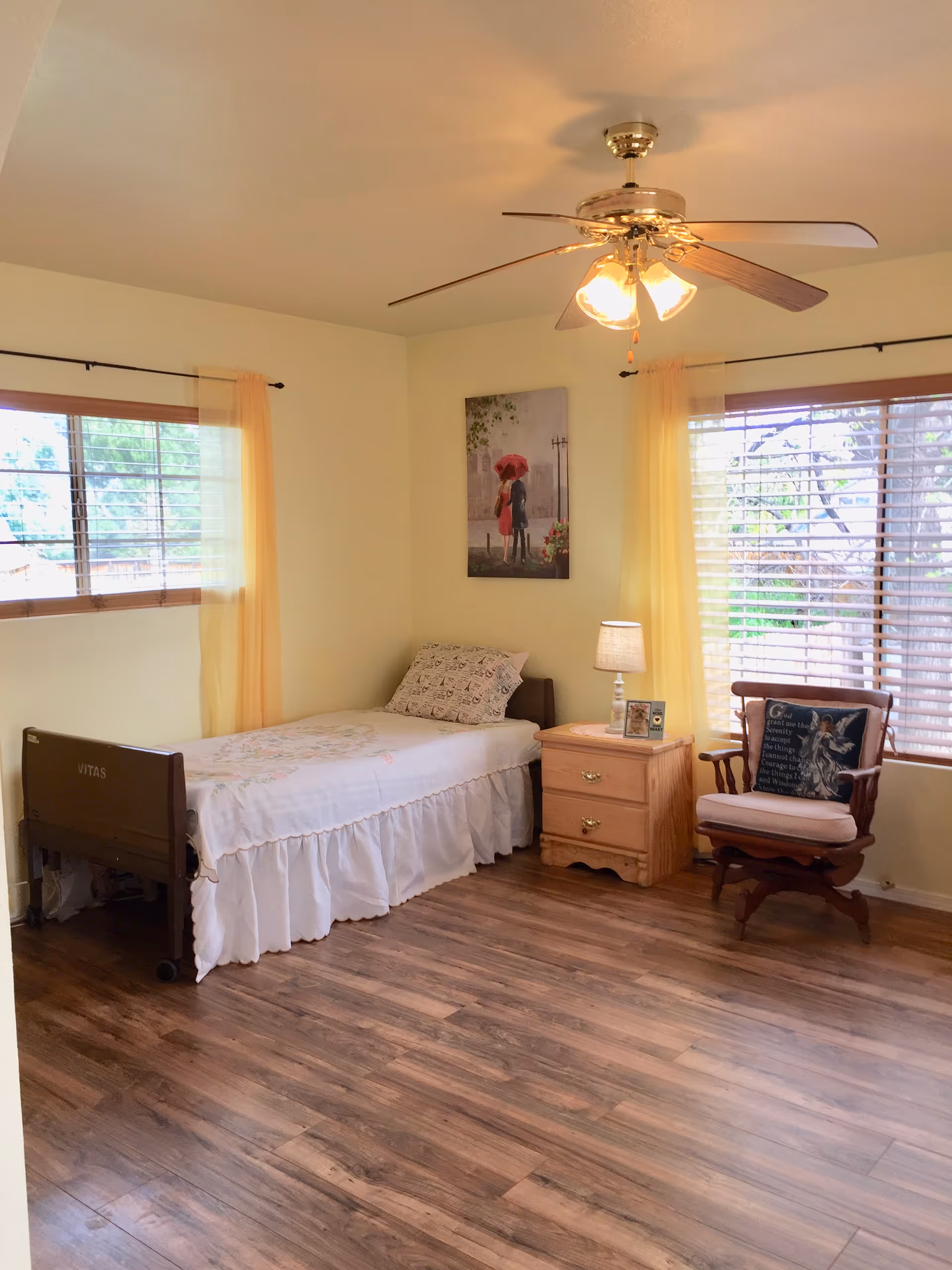 A cozy bedroom in a senior living facility with a bed, nightstand, and large windows.