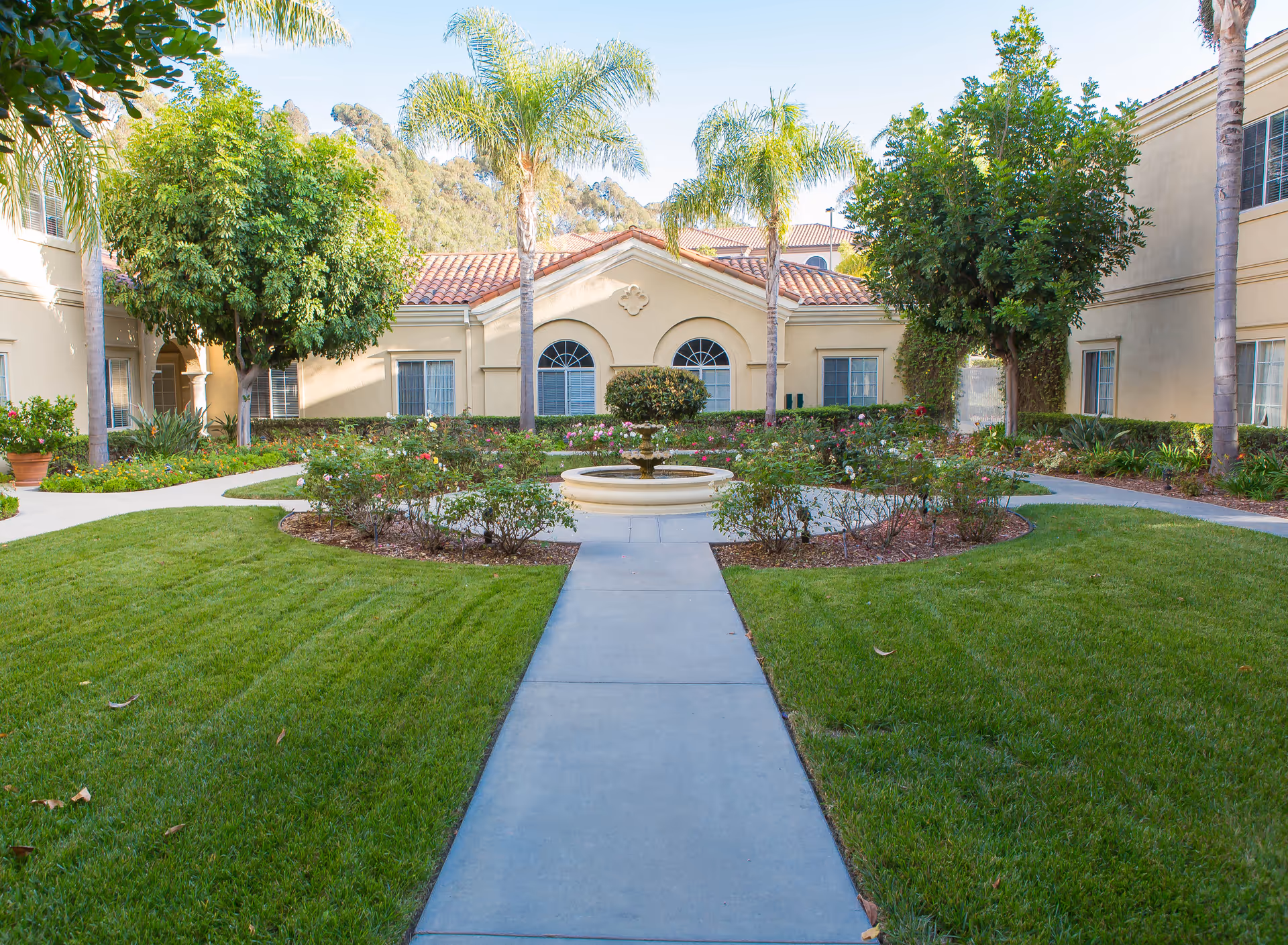 Central courtyard of Bayshire Carlsbad featuring a round fountain, green lawn, and surrounding beige buildings.