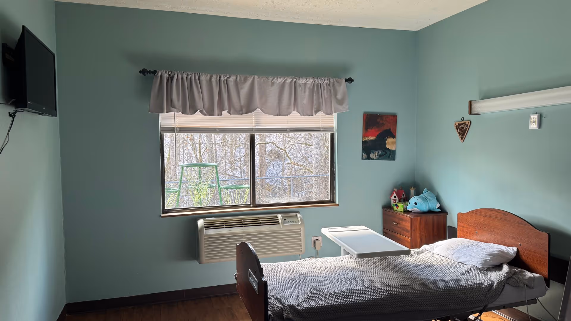 A single resident bed with an overbed table in a teal bedroom of a senior facility.
