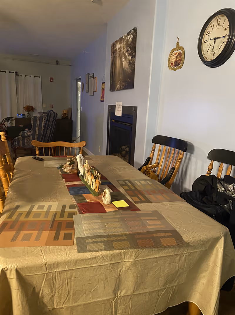 A dining room with a rectangular table covered with a beige tablecloth and a colorful runner in the center. There are six wooden chairs around the table, some with black backs and some natural wood color. On the table, there are placemats, a remote control, and decorative items including a sign that says 'Friends'. The room has light blue walls, a wall clock, a framed picture, and a fireplace. In the background, there is a living area with armchairs and curtains covering a window.