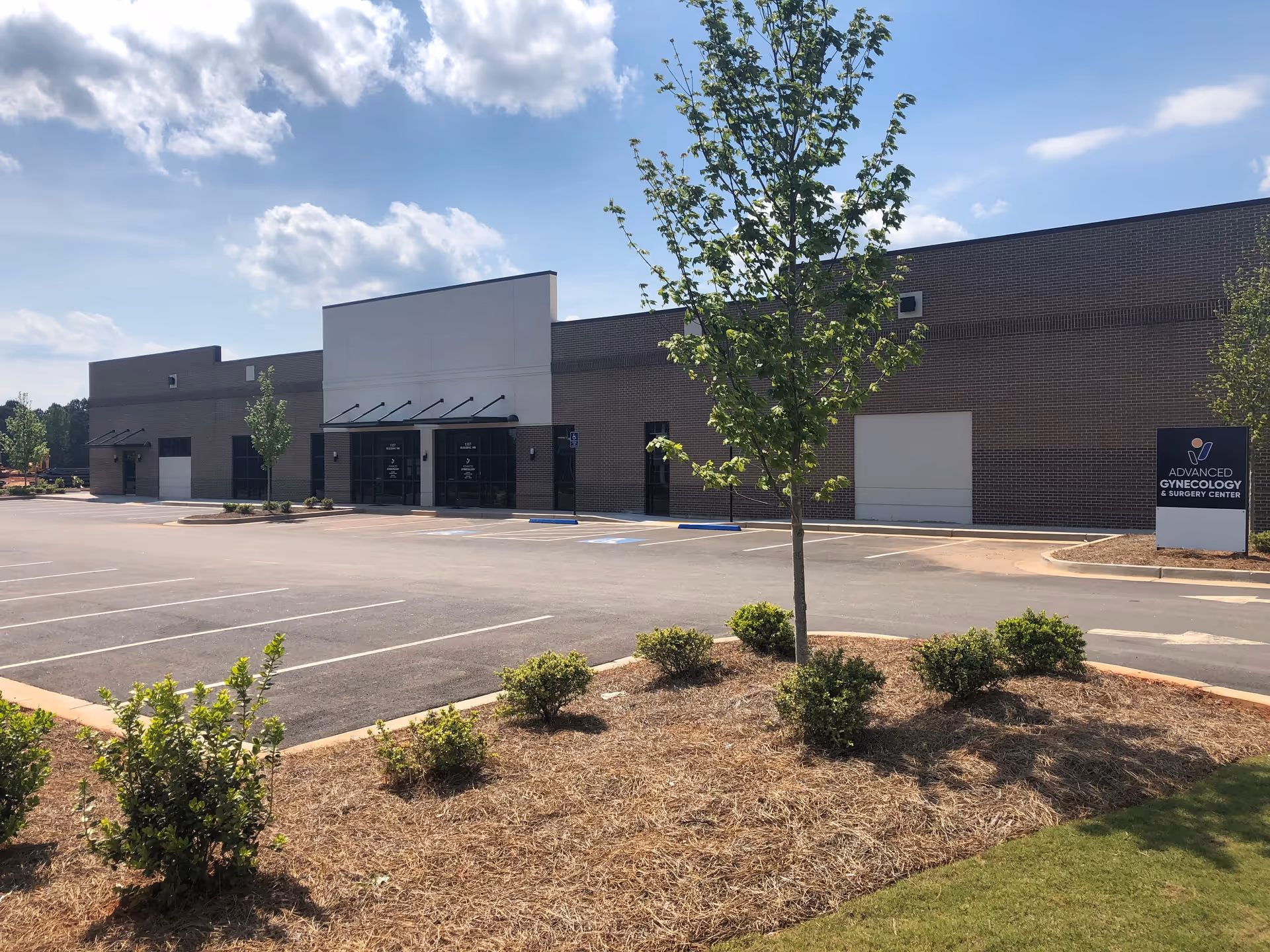 Exterior view of a brick medical building with a sign, parking lot, and trees.